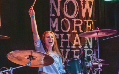 Savannah Tweedt behind her drum kit, lifting one arm triumphantly in the air